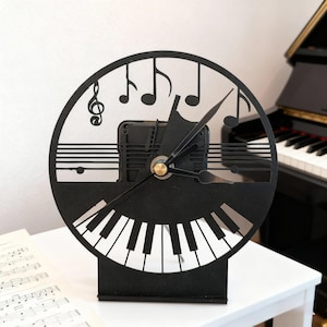 Puede incluir: Reloj negro cortado con láser con un tema musical. El reloj presenta un teclado de piano en la parte inferior, notas musicales y una clave de sol en la parte superior, y un centro dorado. El reloj está sobre una superficie blanca.