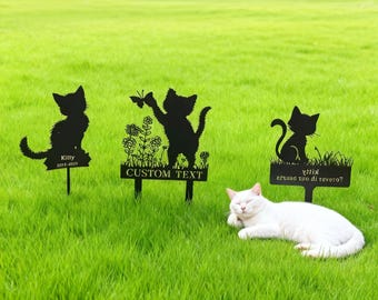 May include: Black metal cat garden stakes with various designs, including a cat silhouette, a cat with a butterfly, and a cat sitting in grass. One stake reads "Kitty 2015-2035", another reads "CUSTOM TEXT". A white cat rests on the green grass.