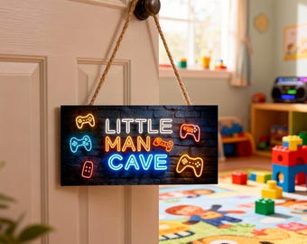 Op de afbeelding: Een rechthoekig houten bord met de woorden "LITTLE MAN CAVE" in neonstijl letters, samen met game controller iconen. Het bord is zwart met een baksteenachtige textuur en hangt aan een witte deur met een touw.