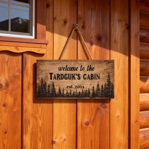 May include: A rectangular wooden sign with the text "welcome to the TARDGUK'S CABIN" and "est. 2004" in black lettering. The sign features a silhouette of pine trees along the bottom edge and is suspended by a rope.