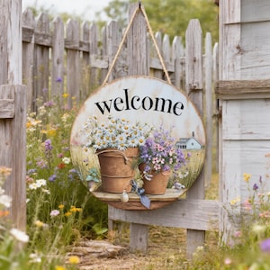 May include: Circular wooden welcome sign with a painted floral design. The sign displays the word "welcome" in black script. Two metal buckets filled with daisies and lavender are shown. The sign hangs from a wooden fence with a rope.