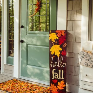 Könnte beinhalten: Ein Holzschild mit den Worten "hello fall" und bunten Blätterillustrationen. Das Schild steht auf einer Veranda neben einer Fußmatte mit Blättermuster. Ein Kranz hängt an der grünen Tür.