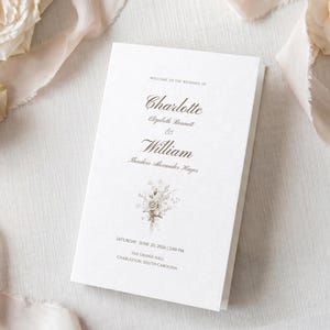 May include: A folded white wedding program with elegant gold lettering. The program reads "Welcome to the Wedding of Charlotte & William" with the date and location at the bottom. A floral illustration is centered.