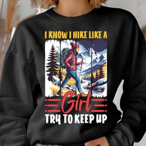 Op de afbeelding: Zwarte sweatshirt met een afbeelding van een wandelaar in een roze jas en blauwe jeans, wandelend in de bergen. De tekst luidt "I KNOW I HIKE LIKE A GIRL TRY TO KEEP UP."