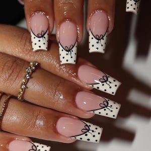 May include: A set of manicured nails with a French tip design. The nails feature a light pink base, white tips with black polka dots, and a black bow design. The nails are long and square-shaped.