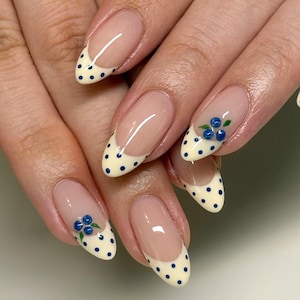 May include: Close-up of manicured nails with a French tip design. The tips are cream-coloured with navy blue polka dots. Some nails feature a small blueberry design with green leaves. The nails are oval-shaped and have a clear base.