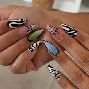 May include: A hand displaying a set of long, almond-shaped acrylic nails. The nails feature various designs, including black and white zebra stripes, black polka dots, and solid colors like pink, green, and blue, each with gold accents.