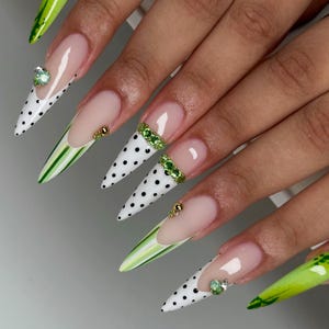 May include: Long stiletto-shaped artificial nails featuring a mix of designs including white tips with black polka dots, green and white vertical stripes, and solid lime green tips with abstract green patterns, accented with small green rhinestones.
