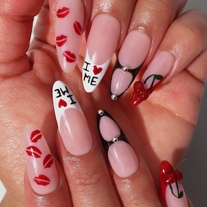 May include: A hand with long, almond-shaped nails. The nails feature a variety of designs, including red lips, the text "I ❤️ ME", and cherry accents. Some nails have a French tip design with black and silver embellishments.