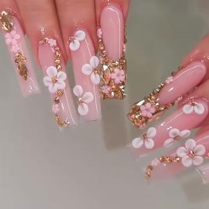 Puede incluir: Uñas artificiales largas de forma cuadrada con una base rosa translúcida, decoradas con acentos florales blancos en tres dimensiones, pequeñas flores rosas y adornos metálicos dorados. Los dedos están adornados con varios anillos dorados.