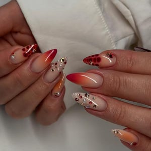Puede incluir: Uñas largas en forma de almendra con un diseño degradado de nude a rojo. Algunas uñas presentan adornos rojos y blancos, mientras que otras tienen un patrón moteado o pequeños acentos de perlas. Uñas bien cuidadas y pulidas.