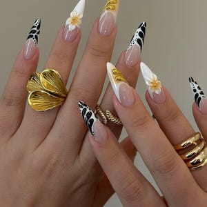 May include: A close-up of manicured nails with a variety of designs. Some nails feature a white base with black abstract patterns, while others have a nude base with white tips and gold accents. One nail has a 3D flower.