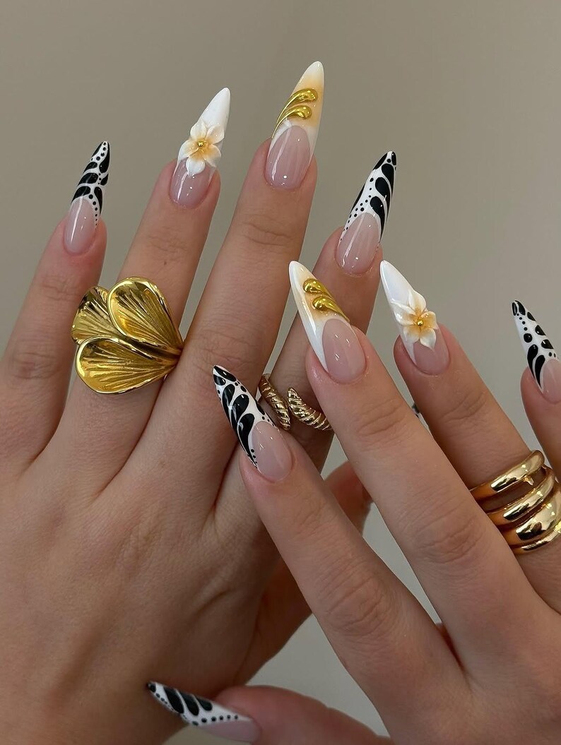 May include: A close-up of manicured nails with a variety of designs. Some nails feature a white base with black abstract patterns, while others have a nude base with white tips and gold accents. One nail has a 3D flower.