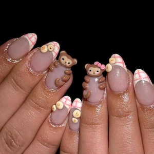 May include: Nail art featuring a nude base with pink and white plaid tips. The design includes three-dimensional brown monkey figures, one with a small pink bow, and tiny yellow banana accents applied to the surface of the manicured fingernails.