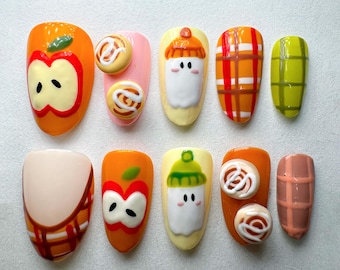 May include: A set of ten artificial nails with various designs. The designs include an apple, a cinnamon roll, a ghost wearing a hat, plaid patterns, and a checkered pattern. The nails are painted in orange, pink, yellow, and brown.