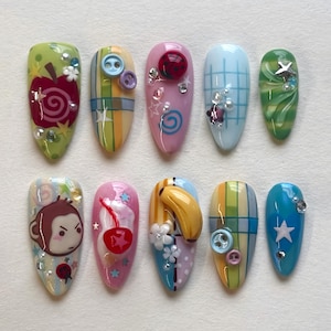 May include: A collection of ten almond-shaped artificial nails, each uniquely decorated. Designs feature an apple, monkey, cherry, banana, buttons, stars, and swirls. The colour palette includes green, pink, blue, yellow, and white, with various embellishments.