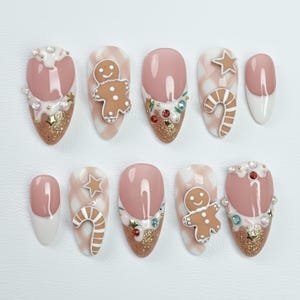 May include: A set of ten almond-shaped, press-on nails with a festive holiday theme. Designs include gingerbread men, candy canes, stars, and glitter accents in shades of pink, white, and gold. Some nails feature a French tip design.