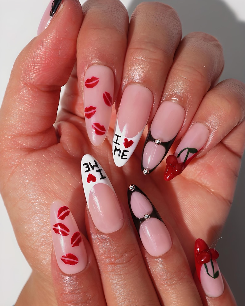 May include: A hand with long, almond-shaped nails. The nails feature a variety of designs, including red lips, the text "I ❤️ ME", and cherry accents. Some nails have a French tip design with black and silver embellishments.