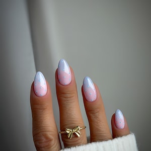 May include: Oval nails with a pink to light blue gradient, decorated with small blue polka dots. A gold bow ring is visible. The nails are neatly manicured and fashionable. The overall look is delicate and stylish.