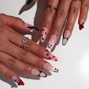 May include: A set of long, almond-shaped acrylic nails. Some nails feature a French tip design with red lip prints, while others have black tips with a jewel accent. The phrase "I ❤️ ME" is written on two nails.