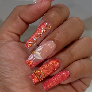 May include: Long, square-shaped artificial nails featuring a vibrant coral-orange glitter finish. One nail displays a white 3D floral accent, while others are adorned with gold-toned metal charms and small, sparkling orange rhinestones.