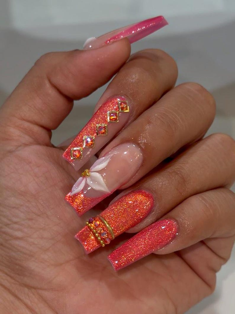 May include: Long, square-shaped artificial nails featuring a vibrant coral-orange glitter finish. One nail displays a white 3D floral accent, while others are adorned with gold-toned metal charms and small, sparkling orange rhinestones.