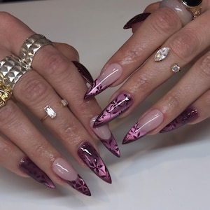 Peut inclure: Ongles longs en forme de stylet avec une manucure française aux pointes prune foncé. Les pointes sont ornées d'un motif floral sombre et complexe. Plusieurs bagues dorées avec diverses pierres sont portées sur les doigts.