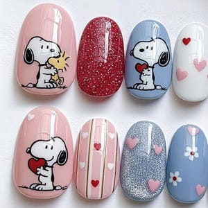 May include: A set of artificial nails with various designs. Some nails feature Snoopy and Woodstock, others have hearts, stripes, and glitter. Colors include pink, red, blue, and white. The nails are oval-shaped and have a glossy finish.