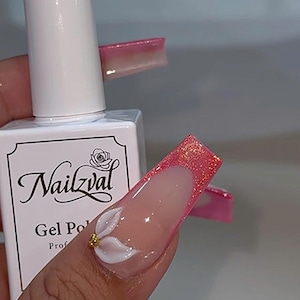 May include: A white bottle of Nailzval gel polish with a black label featuring a rose icon and text. The nail art features long, square-shaped nails with a pink glitter French tip and a three-dimensional white flower accent with a gold center.