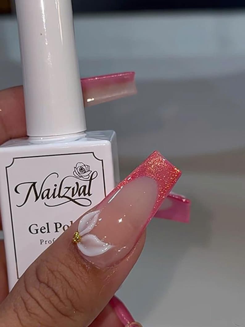 May include: A white bottle of Nailzval gel polish with a black label featuring a rose icon and text. The nail art features long, square-shaped nails with a pink glitter French tip and a three-dimensional white flower accent with a gold center.