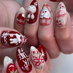 May include: A set of almond-shaped, decorated fingernails. Designs include a white bunny, strawberries, floral patterns, and the text "UR CUTE" on a red background. Accents include silver rhinestones and a gingham pattern.