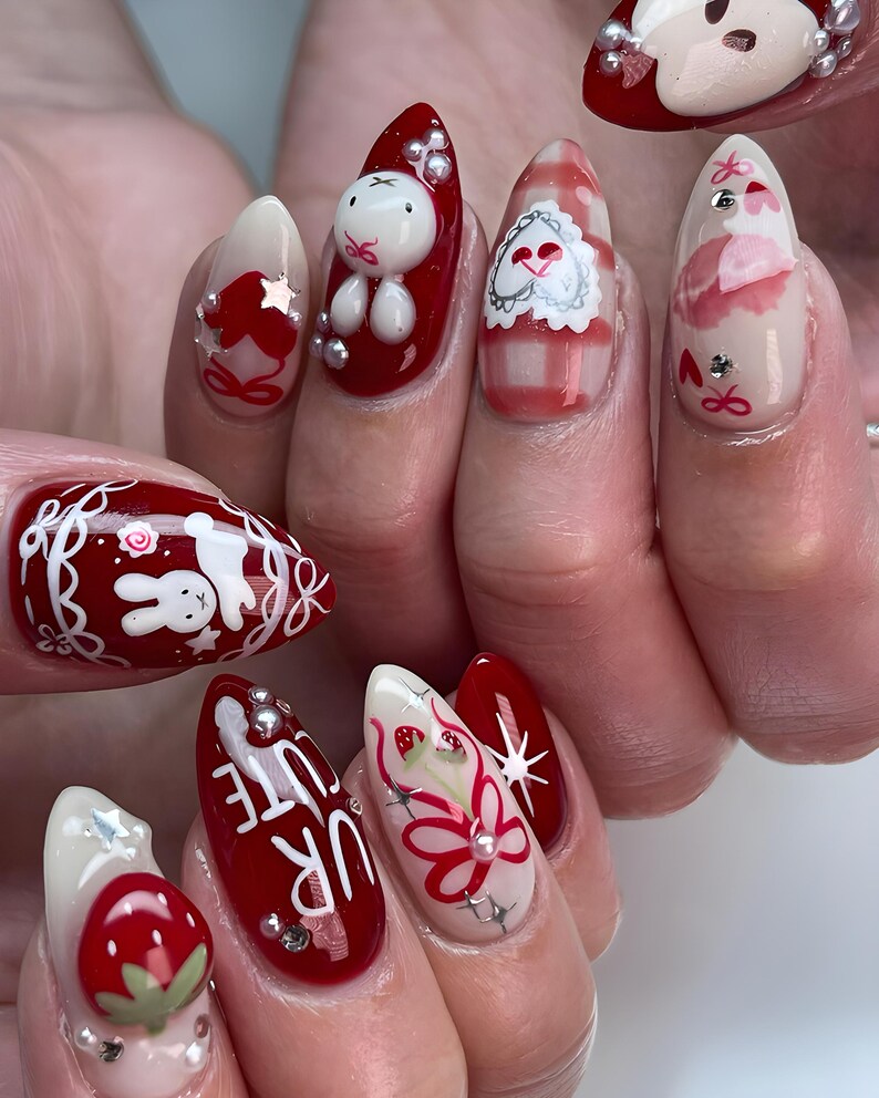 May include: A set of almond-shaped, decorated fingernails. Designs include a white bunny, strawberries, floral patterns, and the text "UR CUTE" on a red background. Accents include silver rhinestones and a gingham pattern.