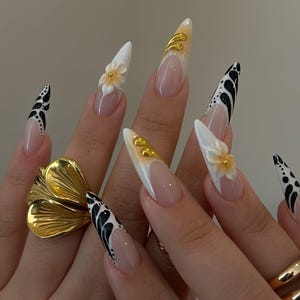 May include: Long, pointed acrylic nails with various designs. Some nails feature a white base with black abstract patterns, while others have a nude base with white tips and gold accents. One nail has a 3D flower. A gold flower ring is also visible.