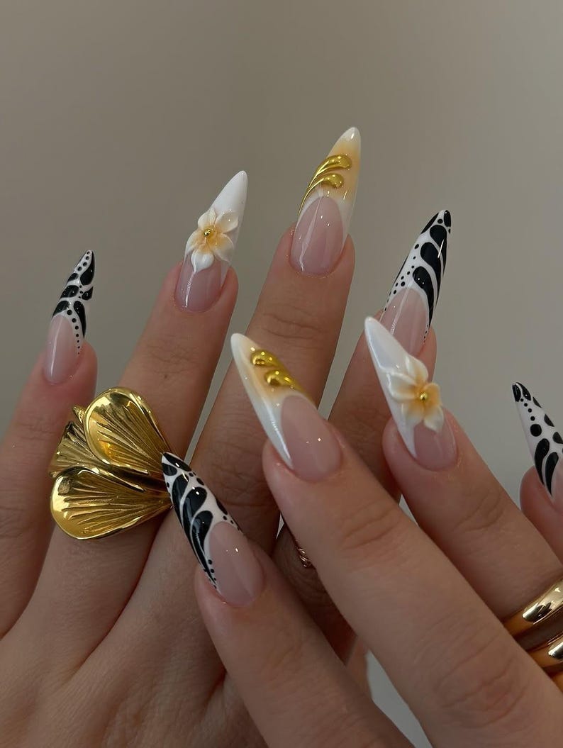 May include: Long, pointed acrylic nails with various designs. Some nails feature a white base with black abstract patterns, while others have a nude base with white tips and gold accents. One nail has a 3D flower. A gold flower ring is also visible.