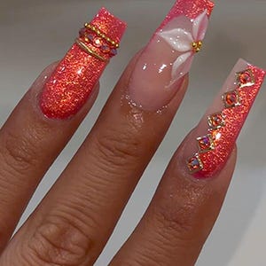 May include: Long square-shaped artificial nails featuring a vibrant coral glitter finish. One nail displays a white 3D flower with a gold center, while the others are accented with gold metal bands and small, square-cut iridescent orange rhinestones.