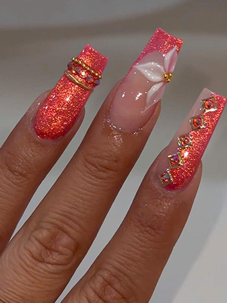 May include: Long square-shaped artificial nails featuring a vibrant coral glitter finish. One nail displays a white 3D flower with a gold center, while the others are accented with gold metal bands and small, square-cut iridescent orange rhinestones.