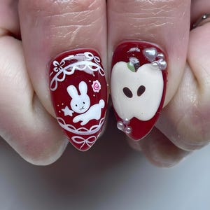 May include: Red nail art featuring a white bunny design with a star and bow, and an apple design with pearl accents. The nails are painted with a glossy red polish, creating a cute and whimsical aesthetic.