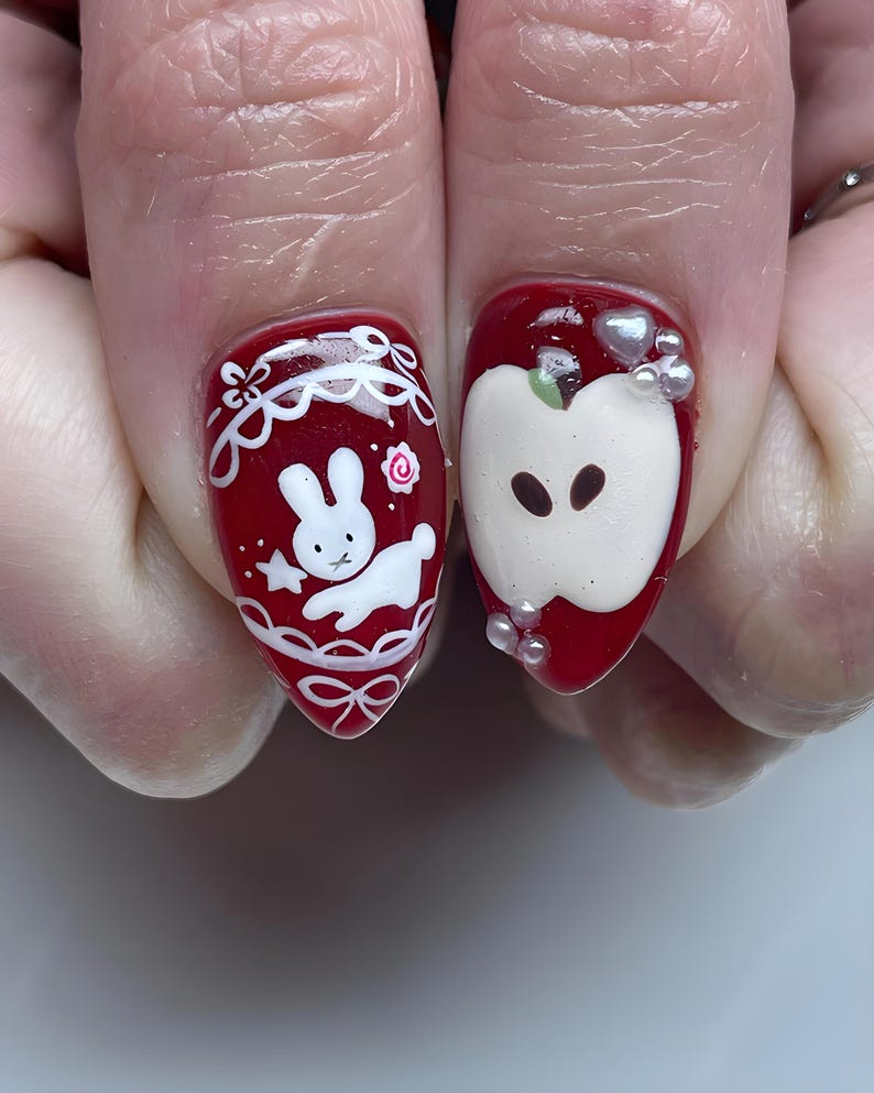 May include: Red nail art featuring a white bunny design with a star and bow, and an apple design with pearl accents. The nails are painted with a glossy red polish, creating a cute and whimsical aesthetic.