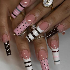 Op de afbeelding: Lange, vierkante kunstnagels met een French manicure-ontwerp. De nagels hebben verschillende patronen, waaronder stippen, strepen en een combinatie van kleuren zoals roze, wit en bruin. Verschillende ringen sieren de vingers.