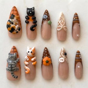 May include: A set of ten almond-shaped press-on nails featuring a nude base with various 3D embellishments including sculpted cats, pumpkins, buttons, and brown and orange patterns. The designs incorporate intricate details like stripes and dots.