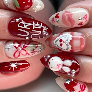 May include: A set of almond-shaped, decorated fingernails. Designs include strawberries, bows, hearts, and a bunny. One nail features the words "UR CUTE". Embellishments include silver stars and small pearls. The nails are painted in shades of red, white, and pink.