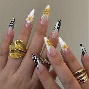 May include: Long, almond-shaped artificial nails with various designs. Some nails are white with gold accents, others feature black and white patterns, and some have small, yellow flower embellishments. Gold rings adorn the fingers.