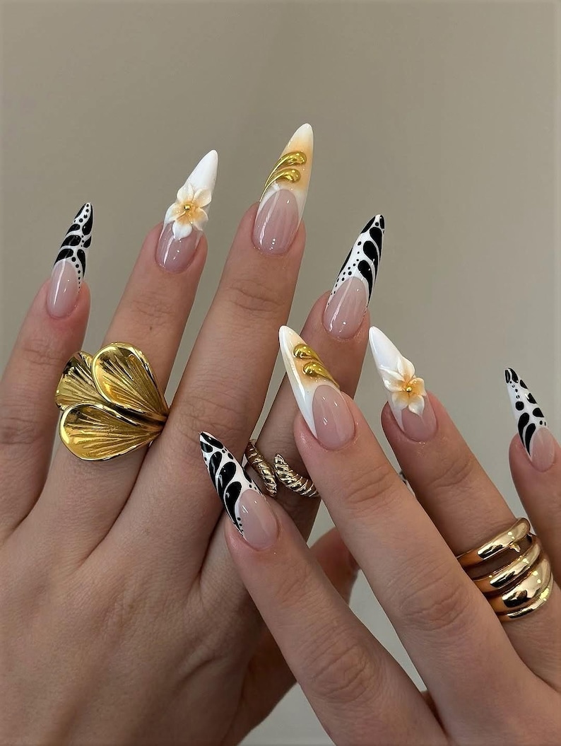 May include: Long, almond-shaped artificial nails with various designs. Some nails are white with gold accents, others feature black and white patterns, and some have small, yellow flower embellishments. Gold rings adorn the fingers.