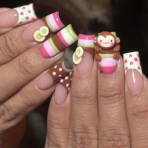 Könnte beinhalten: Lange, geformte Fingernägel mit verschiedenen Designs. Einige Nägel zeigen ein rosa-weißes Polka-Dot-Muster, während andere braune, rosa und grüne Streifen aufweisen. Ein Nagel hat ein 3D-Affen-Design, und mehrere haben bananenförmige Verzierungen.