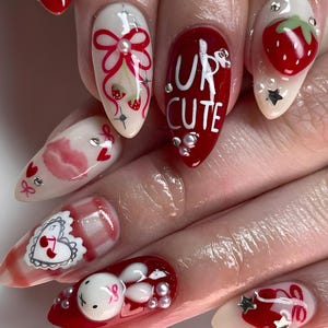 May include: A set of manicured nails with various designs. The nails feature a combination of red, white, and clear polish with embellishments like strawberries, bows, and the text "UR CUTE". Some nails have a glossy finish, while others have a matte appearance.