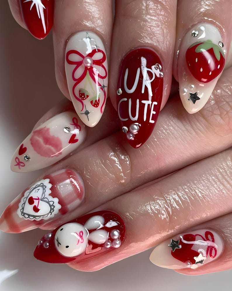 May include: A set of manicured nails with various designs. The nails feature a combination of red, white, and clear polish with embellishments like strawberries, bows, and the text "UR CUTE". Some nails have a glossy finish, while others have a matte appearance.