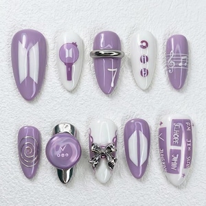 May include: A collection of ten artificial nails in various designs. The nails are in shades of purple and white, with designs including a logo, musical notes, and text. Some nails have silver accents and a bow.