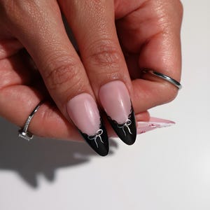 May include: Close-up of manicured nails with a pink base and black tips. The black tips are decorated with a scalloped edge and small silver bow designs. A silver ring with a square stone is visible on the ring finger.