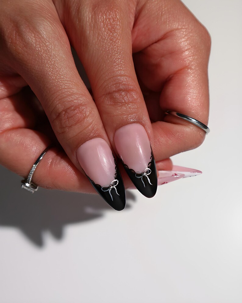 May include: Close-up of manicured nails with a pink base and black tips. The black tips are decorated with a scalloped edge and small silver bow designs. A silver ring with a square stone is visible on the ring finger.