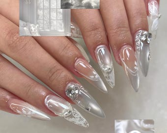 May include: Long, pointed, pearl-white acrylic nails with intricate designs. Some nails feature clear tips, silver glitter, lace, and pearl accents. Small images of a window and a building are in the background.